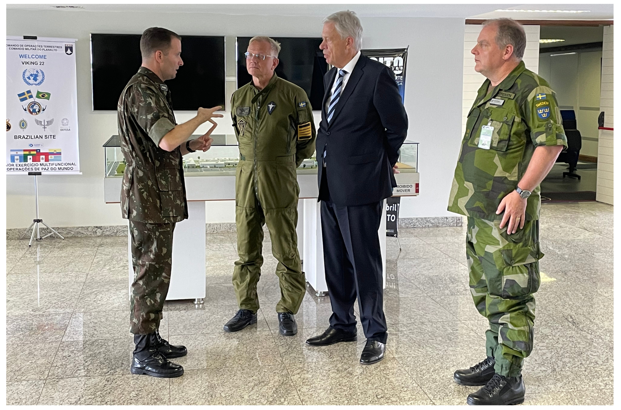 Quatro homens em pé. Três deles à direita escutam um à esquerda. Na ordem, da esquerda para a direita, dois militares, um civil e outro militar.