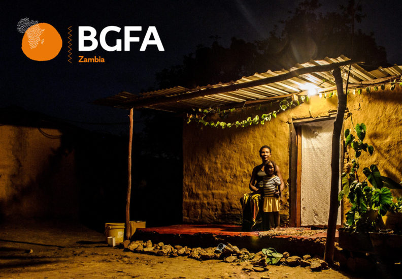 Young girl standing next to her mum outside their house. It is dark outside and a solar powered light bulb is lighting up their veranda.